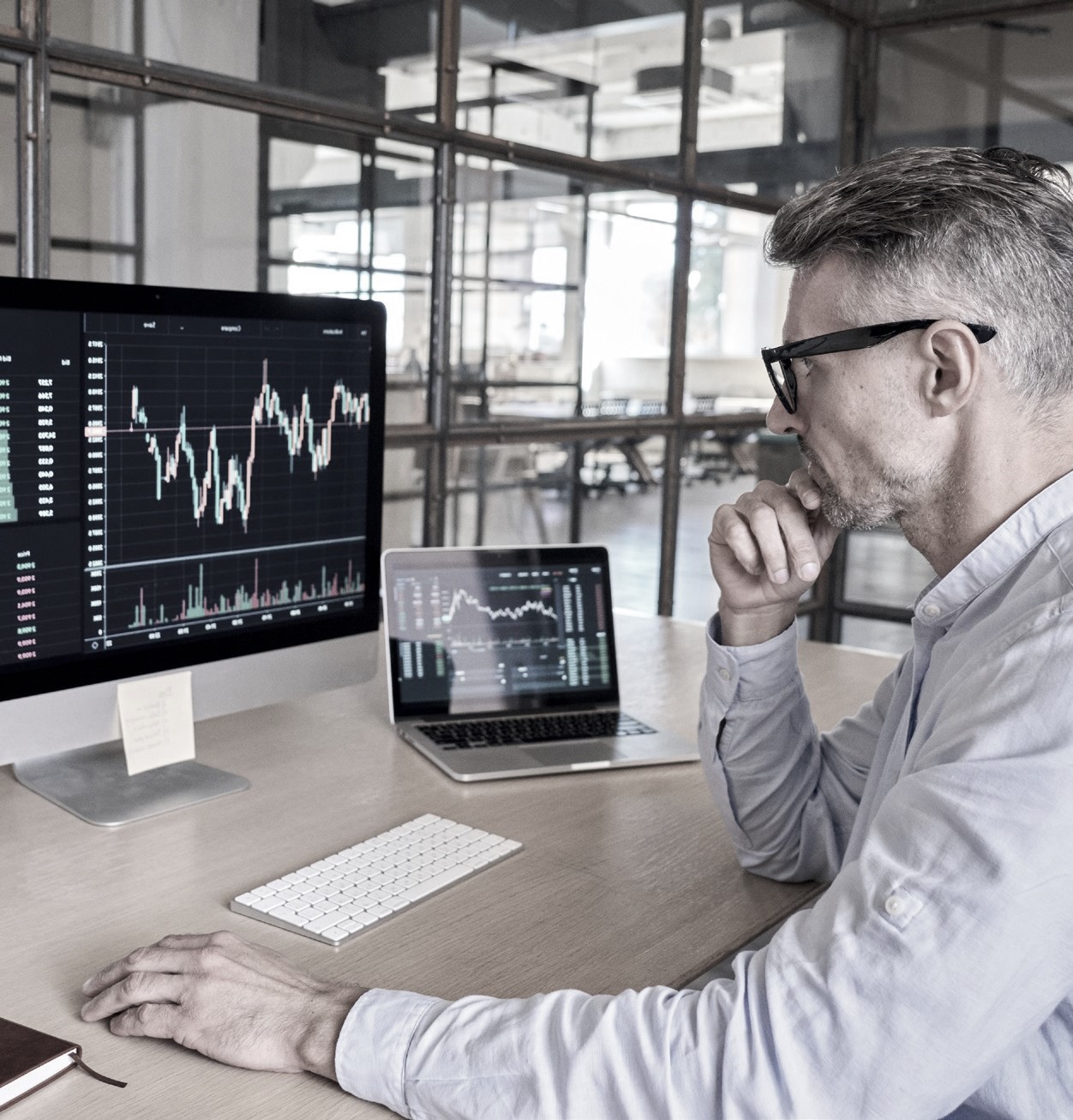 A man wearing glasses sitting at a desk with dual monitors, looking at one of them